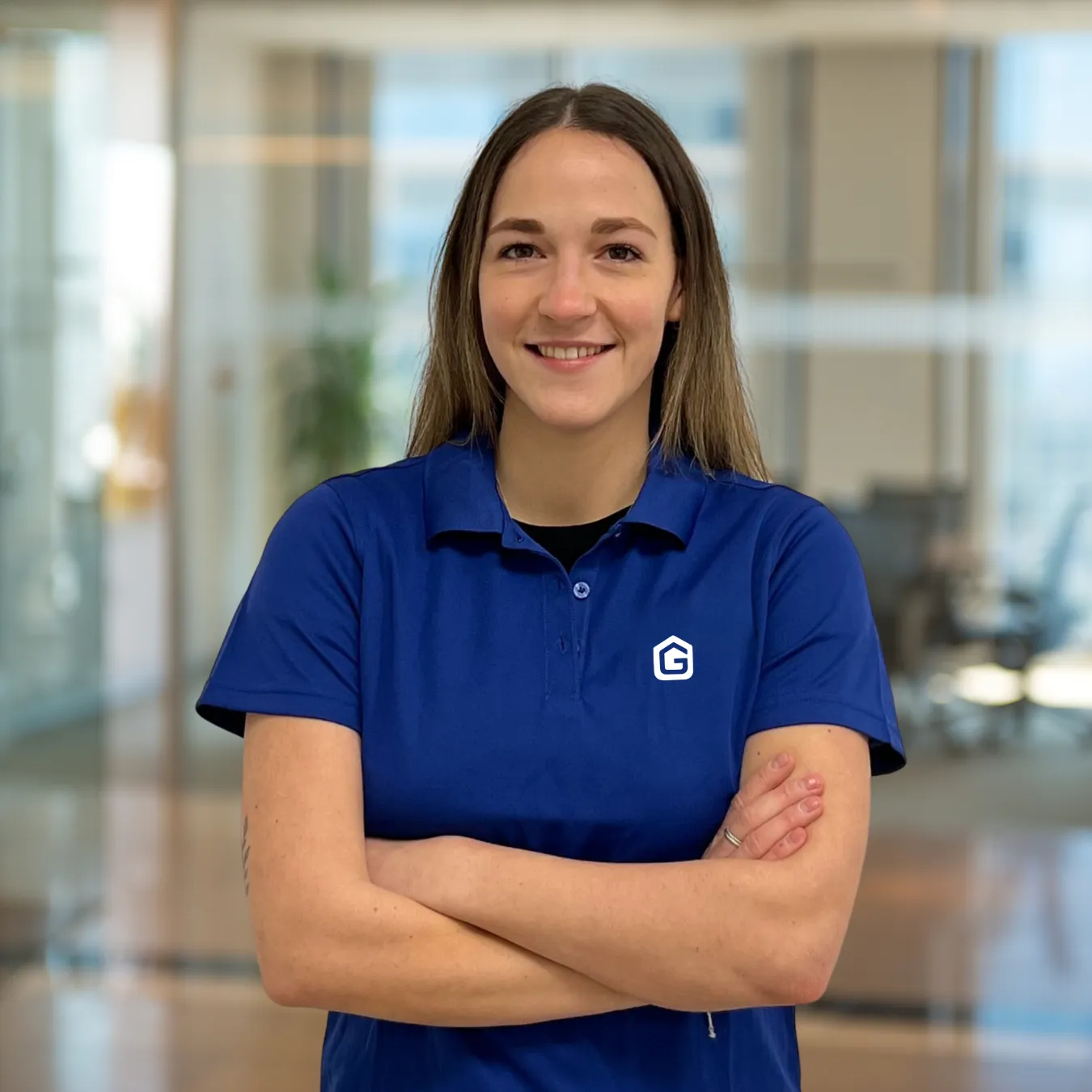 Smiling woman in a blue polo shirt with folded arms standing in a modern office space.