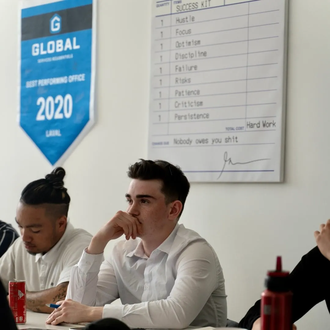Two men sitting at a table in a meeting room with a blue banner that reads 'Global Best Performing Office 2020' and a motivational poster listing traits like Hustle, Focus, and Hard Work.