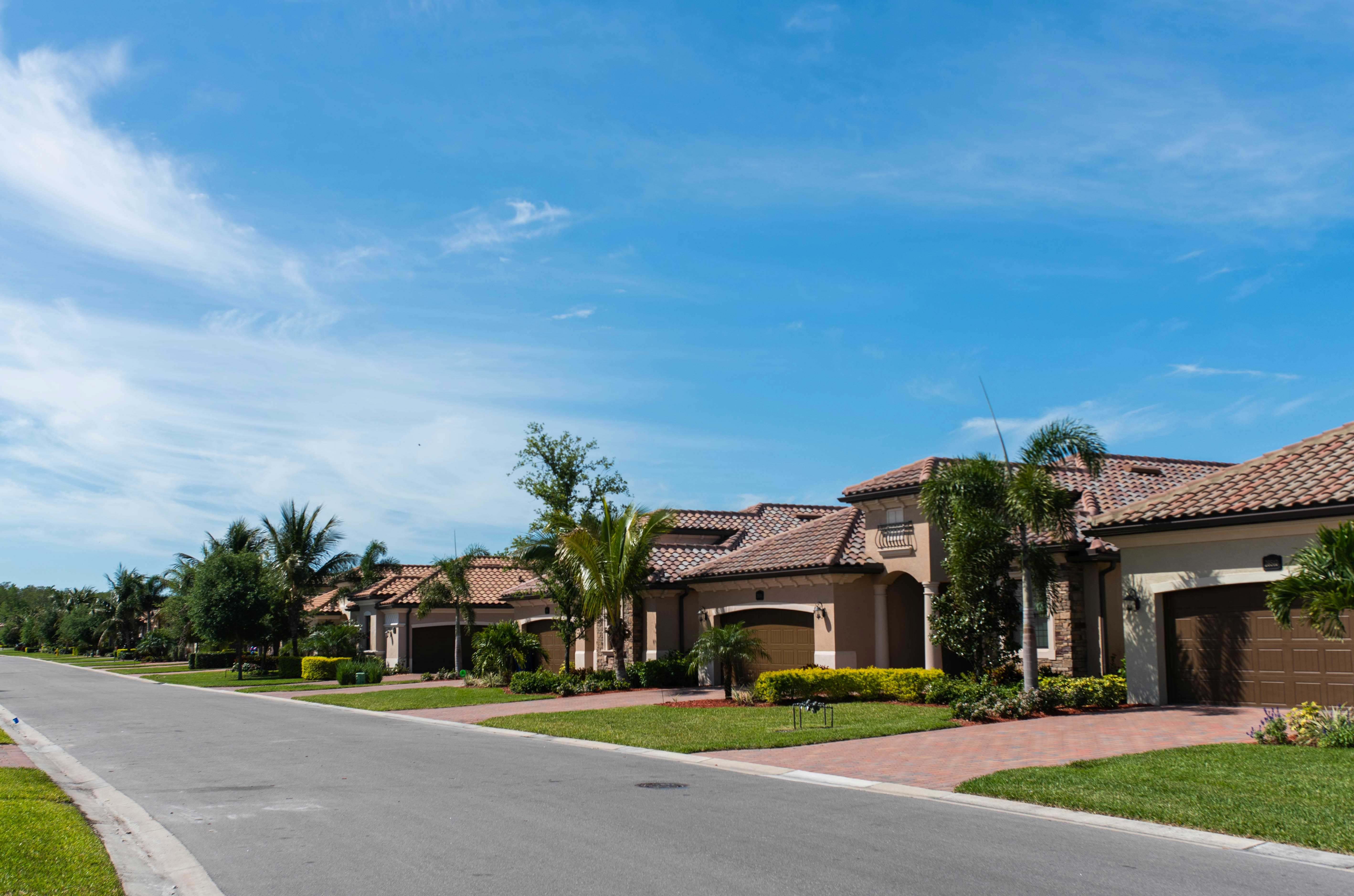 Suburban street with modern single-story houses featuring tiled roofs and palm trees under a partly cloudy blue sky.